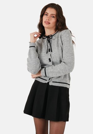 Šedý cardigan s čiernymi akcentmi, ktorý má mašľu na výstrihu, päť gombíkov, rebrované manžety a spodok, kombinovaný s čiernou sukňou.