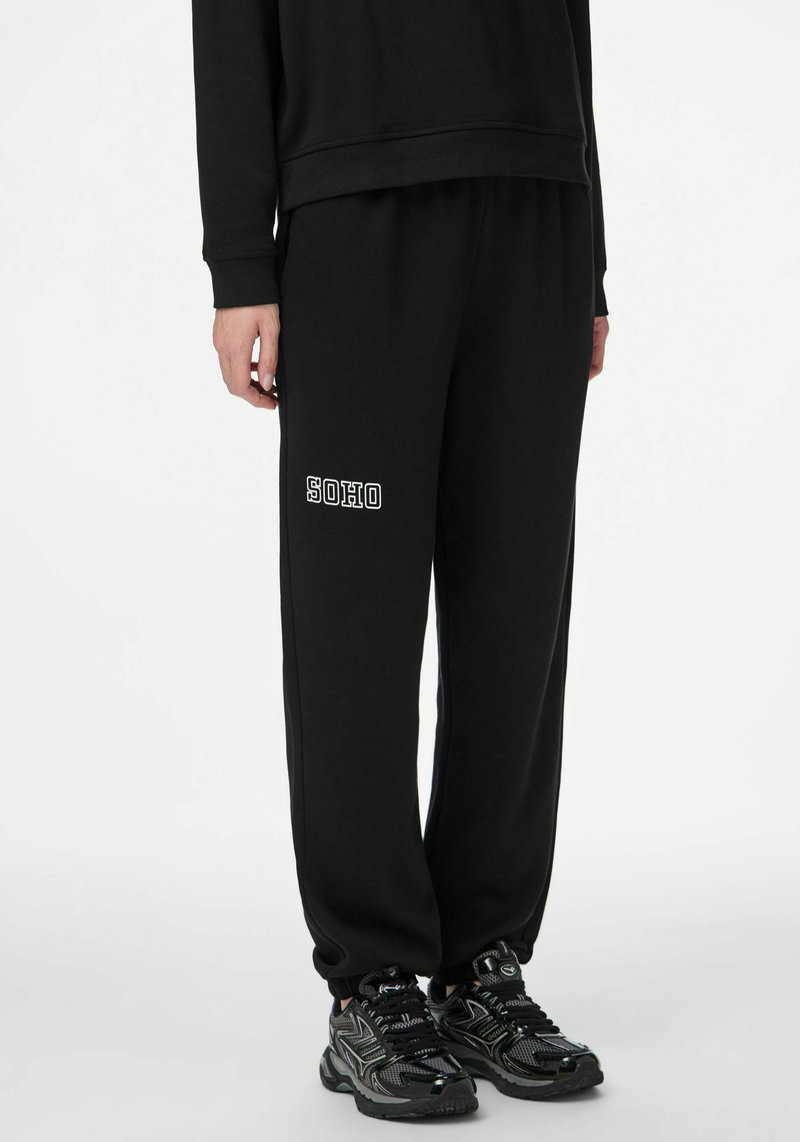 Czarne spodnie dresowe ze ściągaczami przy kostkach, z białym nadrukiem "SOHO" na lewej nogawce, wykonane z miękkiego, teksturowanego materiału.