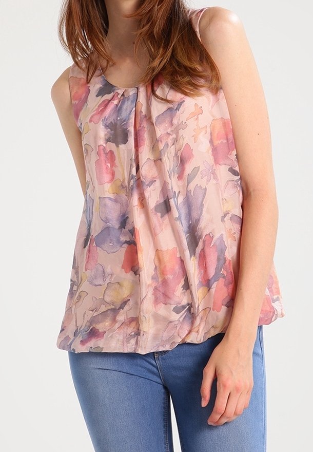 Femme portant un haut sans manches avec un motif floral pastel et un jean bleu, debout devant un fond clair uni.