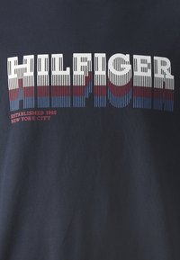 Camiseta de algodón navy con el destacado texto en relieve de rayas blancas y rojas "HILFIGER" y "ESTABLECIDO 1985 NUEVA YORK" en texto más pequeño debajo.