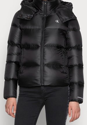 Czarna błyszcząca kurtka puchowa z przednim zamkiem, przyszytym kapturem i logo Calvin Klein na piersi oraz lewym rękawie, noszona z czarnymi spodniami.