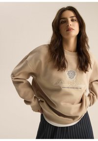 Beżowy sweatshirt z długimi rękawami, ozdobiony haftowanym napisem i emblematem lwa. Miękka tekstura oraz luźny krój. Granatowa spódnica w paski pod spodem.