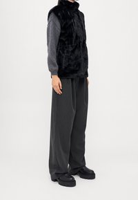 Gilet in pelliccia sintetica nera con collo alto, abbinato a una maglia a maniche lunghe grigia texturizzata e pantaloni larghi a righe. Completa il look un paio di stivali neri robusti.