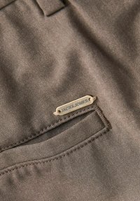 Close-up of brown fabric pants pocket with a small metal label reading "JACK & JONES" sewn above the pocket opening.