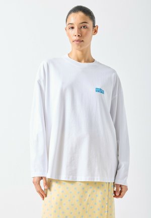 Maglietta a maniche lunghe in cotone bianco con il logo "American Vintage" turchese sul petto a sinistra, abbinata a una gonna gialla a pois.