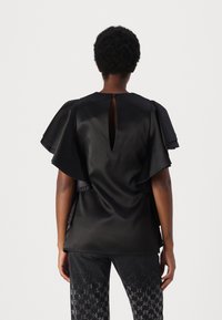 Svart satin topp med volangermar, detail med nøkkelhull bak og glat tekstur. Kombinert med mønstrede mørke bukser med metalliske accenter.