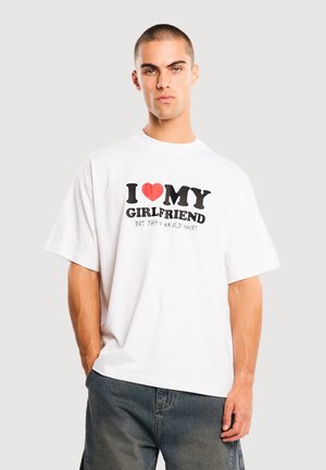 Uomo con capelli corti che indossa una maglietta bianca con la scritta "I [cuore rosso] la mia ragazza ma questa è una maglietta vecchia" e jeans blu scuro, in piedi.