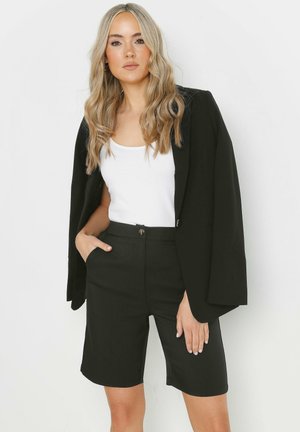 Blazer nero con maniche ampie sopra un top bianco e pantaloni corti neri a vita alta. Texture liscia e vestibilità sartoriale, dettaglio dei bottoni visibile.