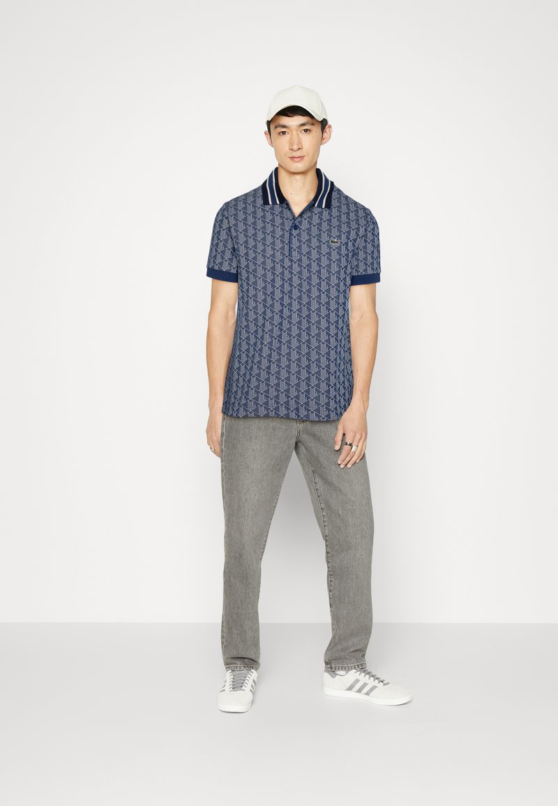 Marineblauwe polo met geometrisch patroon, contrasterende kraag en mouwen, gecombineerd met grijze jeans en witte sneakers. Tegen een eenvoudige achtergrond.