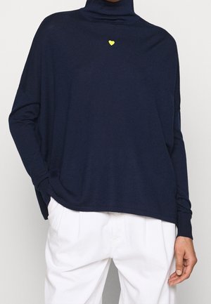 Personne portant un pull à col roulé bleu marine avec un petit cœur jaune en détail et un pantalon blanc, mains partiellement visibles.