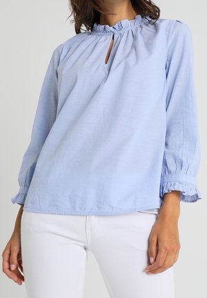 Femme portant un chemisier bleu clair à manches longues avec col et poignets à volants, associé à un pantalon blanc, debout devant un fond uni.