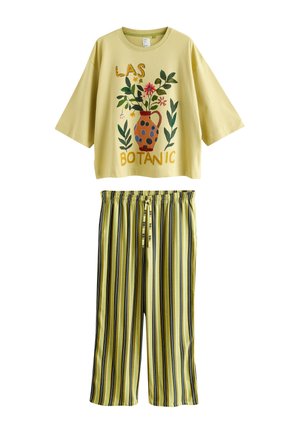 Vrouw in een groen botanisch shirt en groene gestreepte broek, blootsvoets leunend op een houten tafel met een keramische vaas en een fruitmand.
