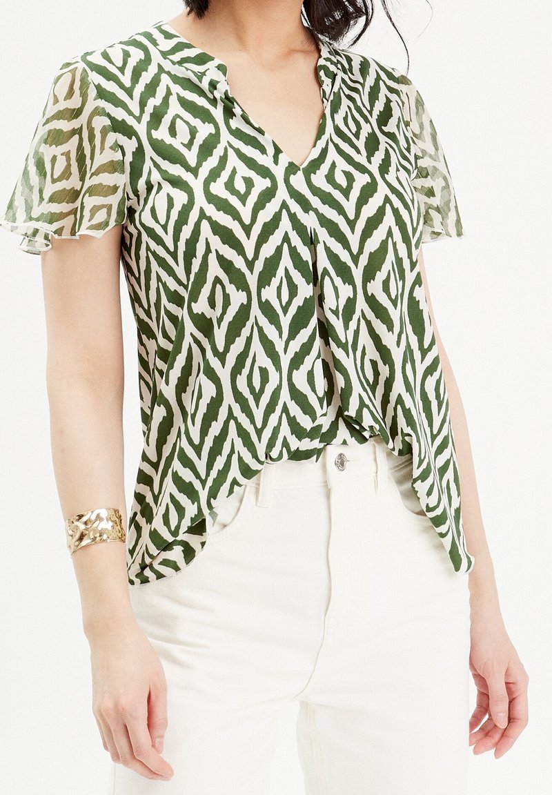 Femme portant un chemisier manches courtes à motifs géométriques verts et blancs et un pantalon blanc taille haute avec un bracelet manchette doré au poignet gauche.