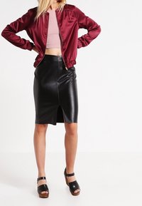 Kobieta nosząca burgundową satynową kurtkę bomber, różowy top cropped, czarną skórzaną spódnicę o ołówkowym kroju z zapięciem z przodu oraz czarne sandały na platformie.