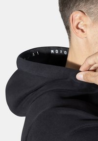 Sudadera negra con capucha que presenta una textura suave, una capucha redondeada y una etiqueta blanca impresa en el interior del cuello. Mano ajustando la capucha.