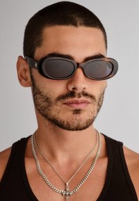Μαύρα και καβουροειδή οβάλ γυαλιά ηλίου με σκούρους φακούς. Συνοδευόμενα από ασημένιες αλυσίδες και φορεμένα με μαύρο κοντομάνικο μπλουζάκι.