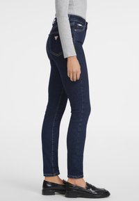 Jeans skinny azul oscuro con una textura de denim suave, cintura alta y bolsillos traseros. Combinados con mocasines negros y una parte superior gris claro.