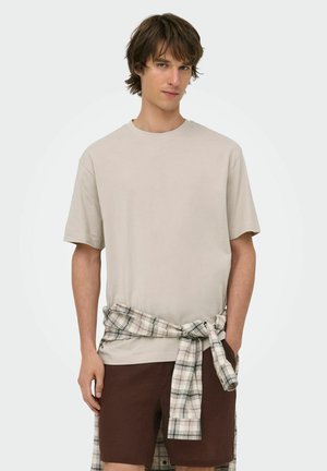 Giovane uomo con capelli castani che indossa una camicia beige a maniche corte, pantaloncini marroni e una camicia a quadri legata intorno alla vita, su uno sfondo uniforme.