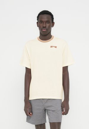 Μπεζ μπλούζα με πορτοκαλί ριμπ λαιμόκοψη, με κεντημένο το «HOUSE OF SUNNY» σε πορτοκαλί. Το μοντέλο φοράει καρό σορτς.