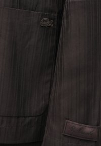 Tissu marron foncé à fines rayures avec une texture douce ; présente un logo Lacoste et une étiquette avec l'inscription embossée "R. Lacoste".