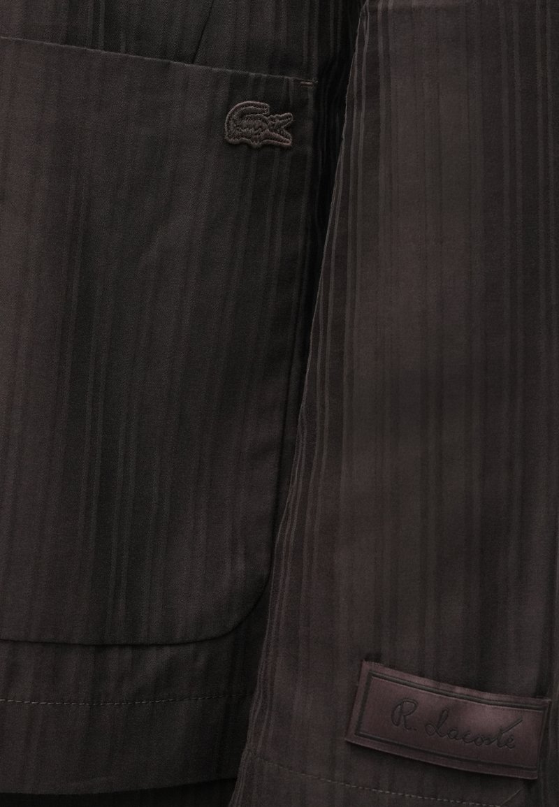 Donkerbruin, stof met krijtstreep en een zachte textuur; voorzien van een Lacoste-logo en een label met "R. Lacoste" in reliëfdruk.