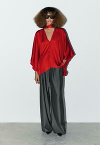 Rode satijnen oversized top met een V-hals en uitsparingen op de schouders, gecombineerd met losse grijze pinstripe broeken. Model draagt donkere zonnebril.