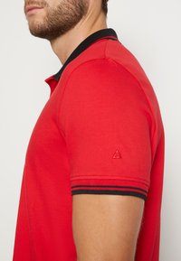 Rotes Poloshirt mit schwarzem geripptem Kragen und Ärmelbündchen, gerippter Struktur und einem kleinen Logodetail am linken Ärmel.