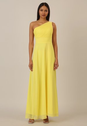 Frau steht frontal und trägt ein ärmelloses, einseitig schulterfreies gelbes Kleid mit plissiertem Oberteil und fließendem Rock, kombiniert mit silbernen High Heels.