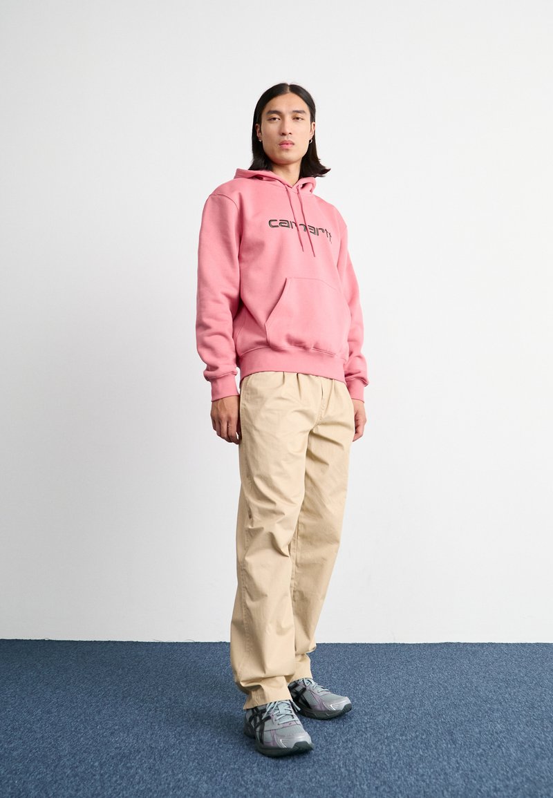 Roze hoodie met een voorzak en logo, gecombineerd met beige broeken en grijze sneakers. De materialen lijken zacht en comfortabel.