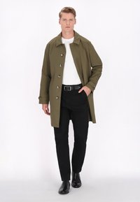 Oliven grøn trenchcoat med krave, seks metallknapper og lange ærmer, parret med sorte bukser og lædersko. Hvid t-shirt under.
