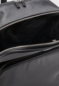 Sac à dos en nylon noir avec fermetures éclair jumelles. L'intérieur présente un motif répétitif du logo "GUESS" sur une doublure noire. Texture lisse.