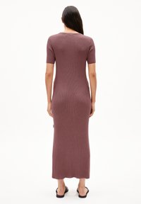 Rippstrick, mauves Strickkleid mit kurzen Ärmeln und einer langen, eng anliegenden Silhouette. Der Saum ist leicht gewellt. Getragen mit schwarzen Schlüpfsandalen.