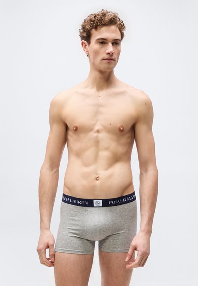 Νεαρός άνδρας με σγουρά μαλλιά που φοράει γκρι εσώρουχα Polo Ralph Lauren, στέκεται μπροστά σε απλό ανοιχτό γκρι φόντο.