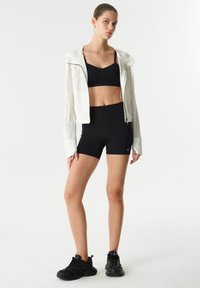 Soutien-gorge de sport noir et short assorti avec un sweat à capuche blanc léger à zip, doté d'un design élégant et d'un tissu respirant. Des baskets noires complètent le look.