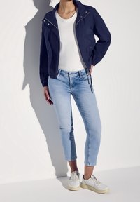 Marinblå lättviktsjacka med silverdragkedja, vit t-shirt, ljusblå jeans och vita sneakers med beige detaljer, alla med avslappnad passform.