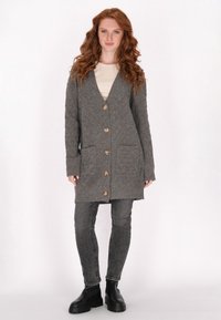 Cardigan grigio a maglia con scollo a V, sei bottoni in legno, due tasche frontali e maniche lunghe. Abbinato a jeans grigio scuro e stivali neri.