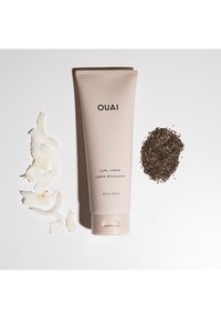Un tube beige étiqueté "OUAI CURL CRÈME" à côté de flocons de noix de coco et d'une pile de graines noires sur un fond blanc.