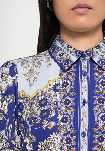 Chemise florale bleue avec un col boutonné, présentant des motifs complexes en bleu, jaune et gris, fabriquée dans un tissu lisse.