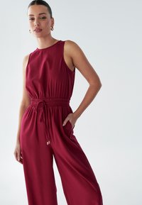Burgundijski brezrokavni kombinezon z elastičnim pasom, stranskimi žepi in sproščenim krojem. Ima gladko teksturo in subtilen sijaj.