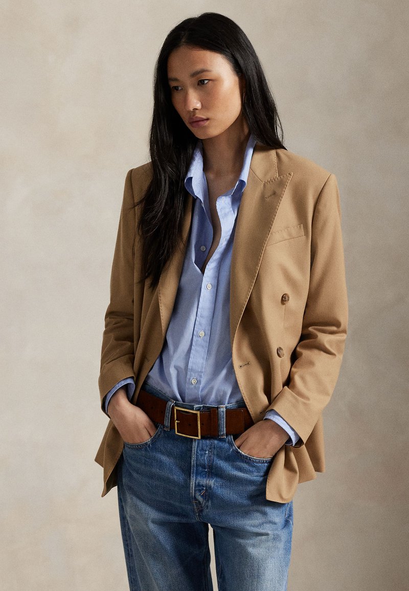 Tanmavý dvojradový blejzer cez svetlomodrú košeľu s gombíkmi, skombinovaný s nohavicami s vysokým pásom v svetlomodrej farbe a hnedým koženým opaskom.