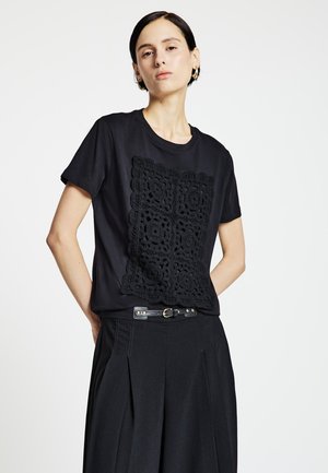 Schwarzes T-Shirt mit kurzen Ärmeln und einem strukturierten, schwarzen Häkelpanel auf der Vorderseite, kombiniert mit hoch taillierten, schwarzen, plissierten Hosen und einem schwarzen Gürtel.