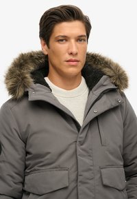 Giacca isolata grigia con cappuccio foderato in pelliccia, dotata di chiusura a zip e due tasche frontali. Indossata sopra un maglione leggero a maglia.
