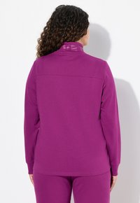 Fialová halenka s dlouhým rukávem a vysokým límcem, která má jemnou texturu a detaily šití. Na zadní straně se nachází logo v tónovém tisku.