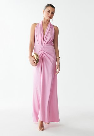 Lange roze halterjurk met plooien en een geplooid middenstuk. Voorzien van een gladde stof en een vloeiende rok. Gecompleteerd met gouden accessoires.