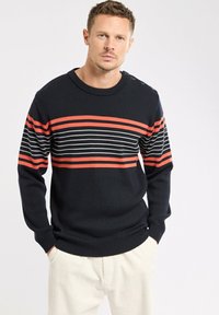 Homme portant un pull noir avec des rayures horizontales rouges et blanches, les mains dans les poches, se tenant contre un fond blanc.