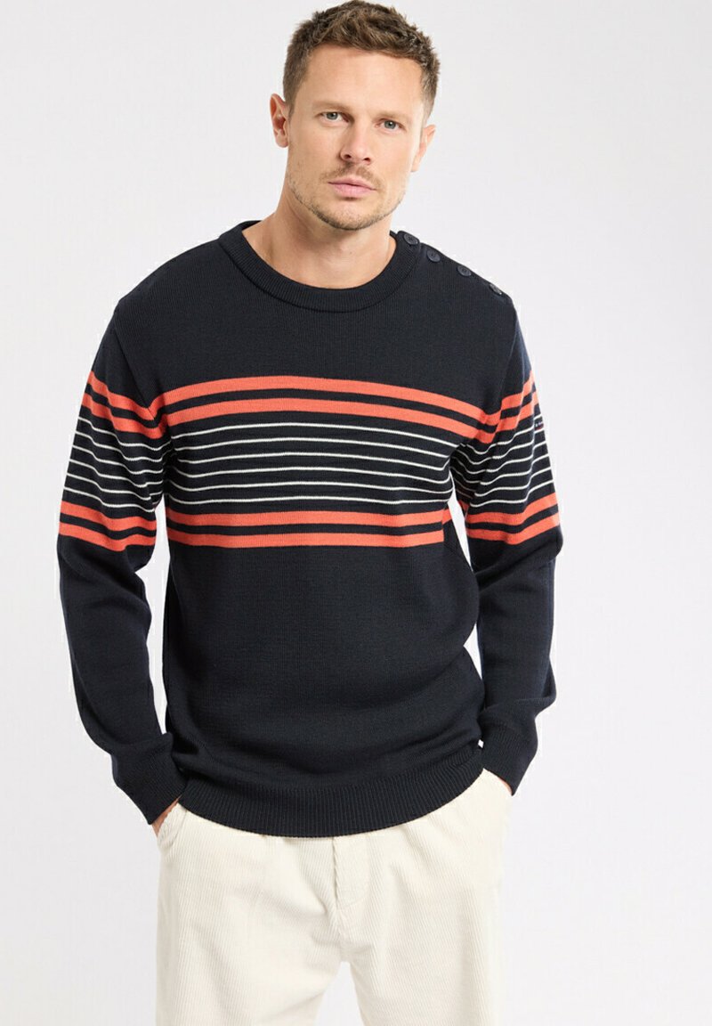 Homme portant un pull noir avec des rayures horizontales rouges et blanches, les mains dans les poches, se tenant contre un fond blanc.