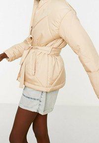 Beige pufferjakke med bælte i taljen, kviltet tekstur og trykknaplukning. Bæret over denimshorts, hvilket viser en afslappet pasform.