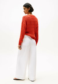 Maglione rosso a maglia con un motivo testurizzato, abbinato a pantaloni bianchi larghi con una tasca sul retro. Si indossano semplici sandali neri.