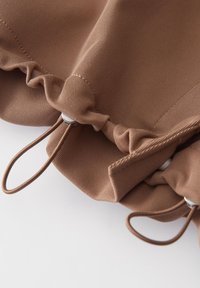 Bruin ribbelstof met geplooide randen, voorzien van decoratieve elastische lussen en zilverkleurige hardware-accenten. Textuur oppervlak met gekruiste details.