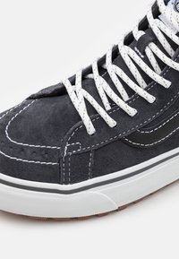 Svart mockaskor med vit sömnad, featuring en vit gummi sula och svarta detaljer på ovansidan. Skosnörena är vita. Strukturerad yta.